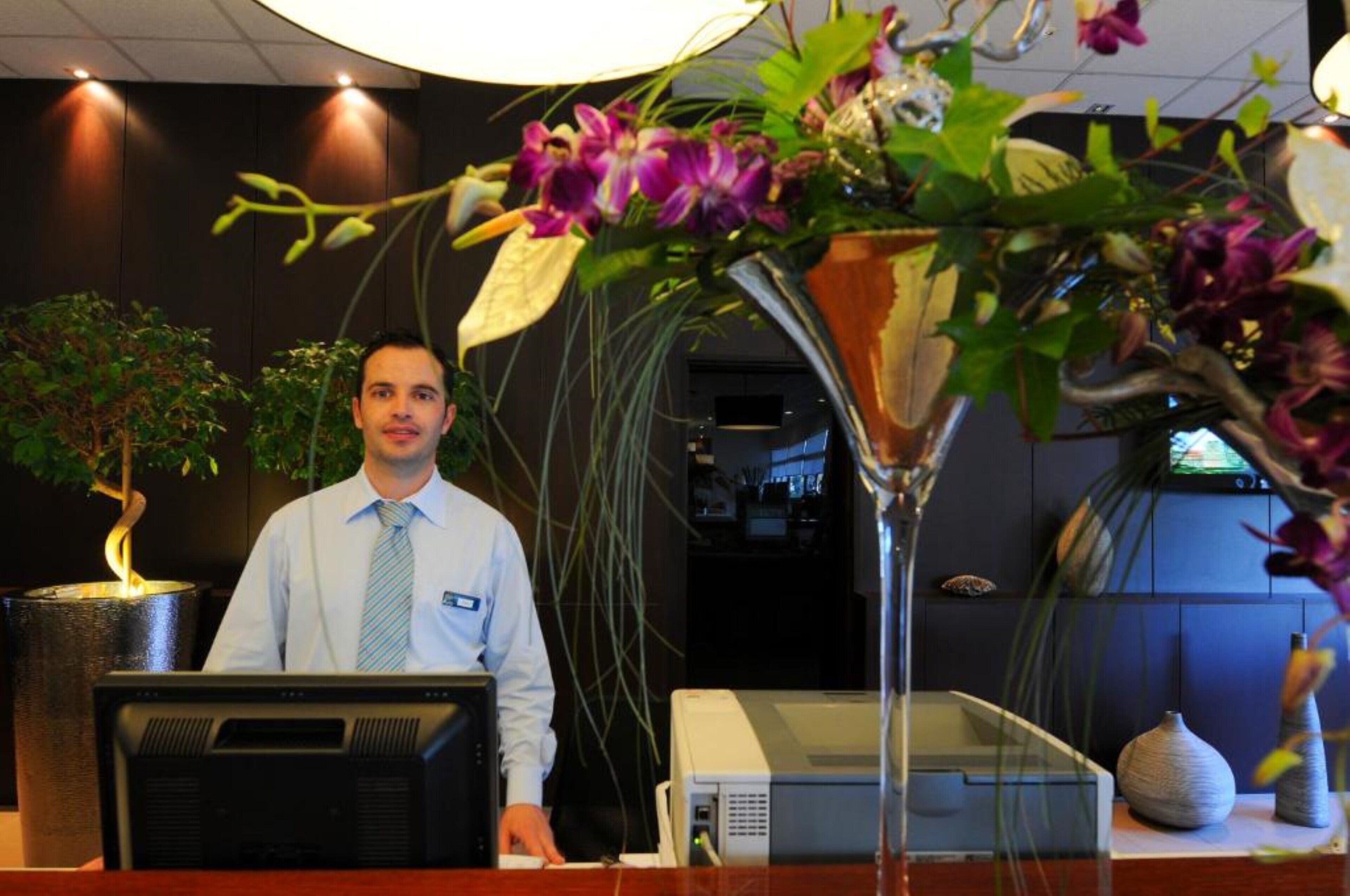 A reception desk with a computer, a large vase with flowers, and a potted plant.