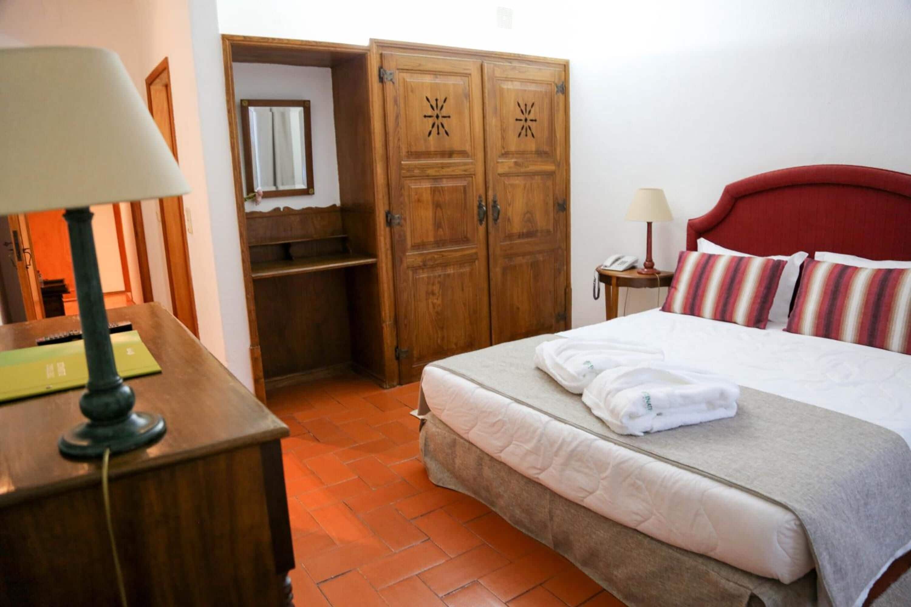 A hotel room with a bed, a desk with a lamp, a wardrobe, and a small bedside table with a lamp.