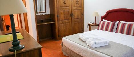 A hotel room with a bed, a desk with a lamp, a wardrobe, and a small bedside table with a lamp.