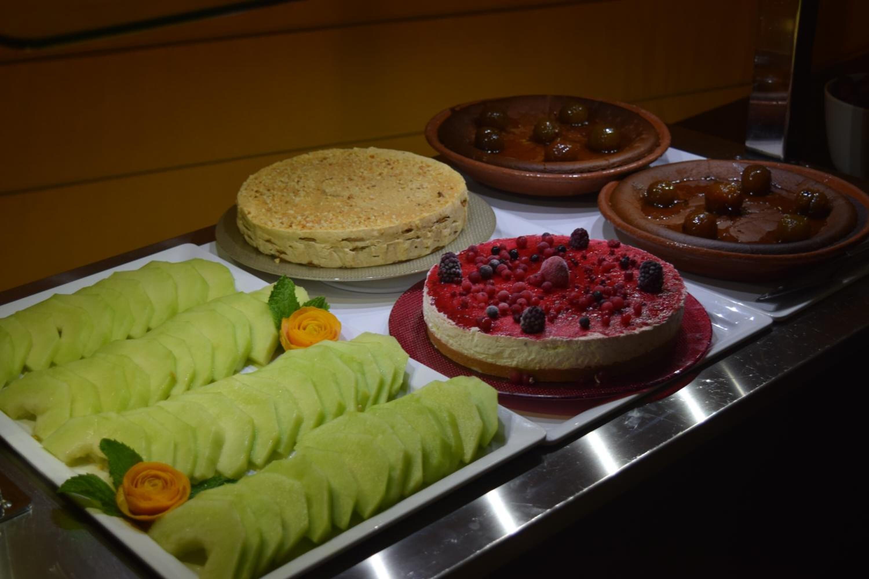 Un bufé con diversos postres, incluyendo un plato de fruta verde, un postre de bayas rojas y dos fuentes marrones con salsa.