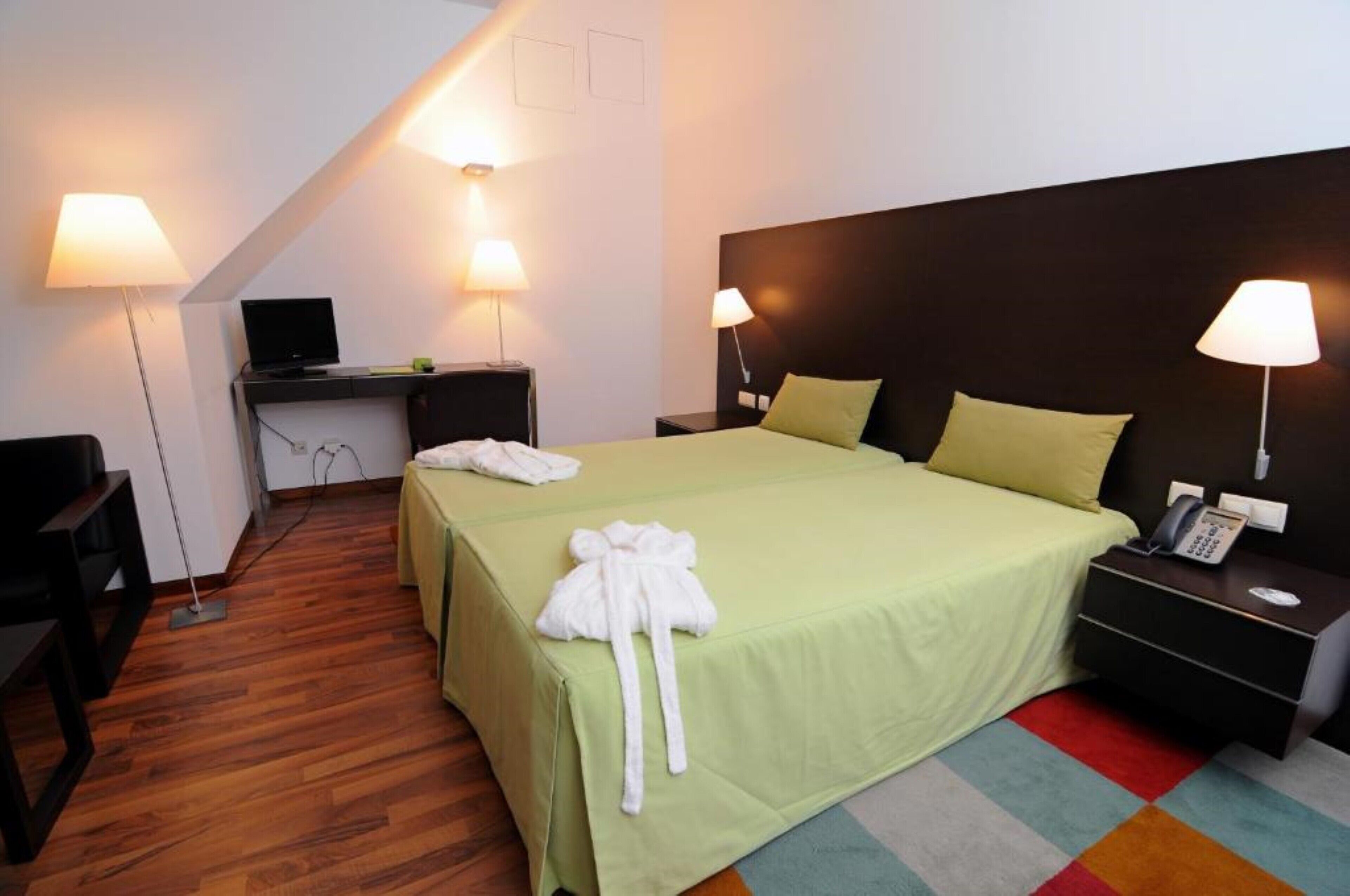 A hotel room with a bed, two bedside tables, a desk with a computer, and two lamps.