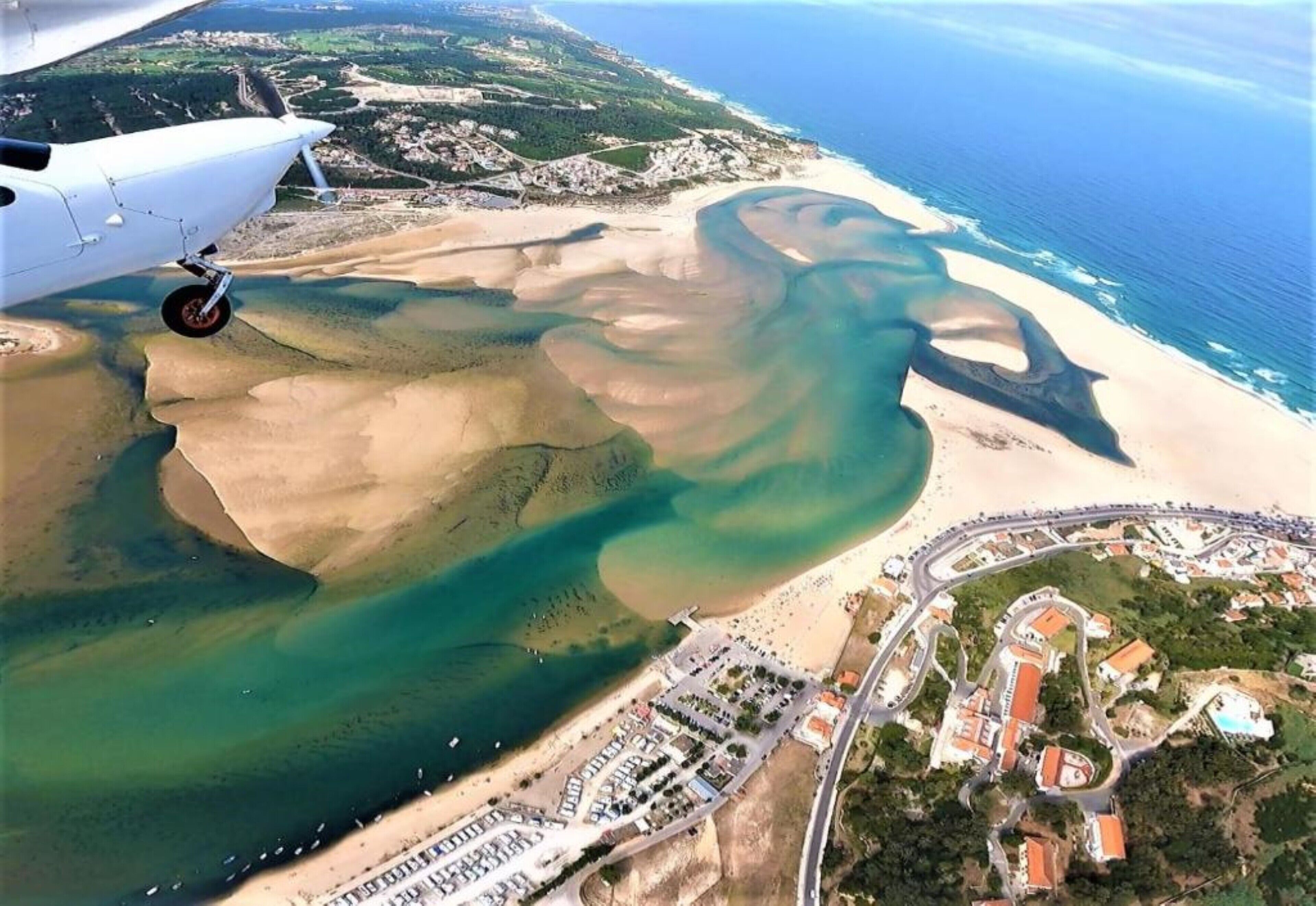 Vista aérea de una zona costera con playa, puerto deportivo y un pequeño pueblo con edificios y caminos.