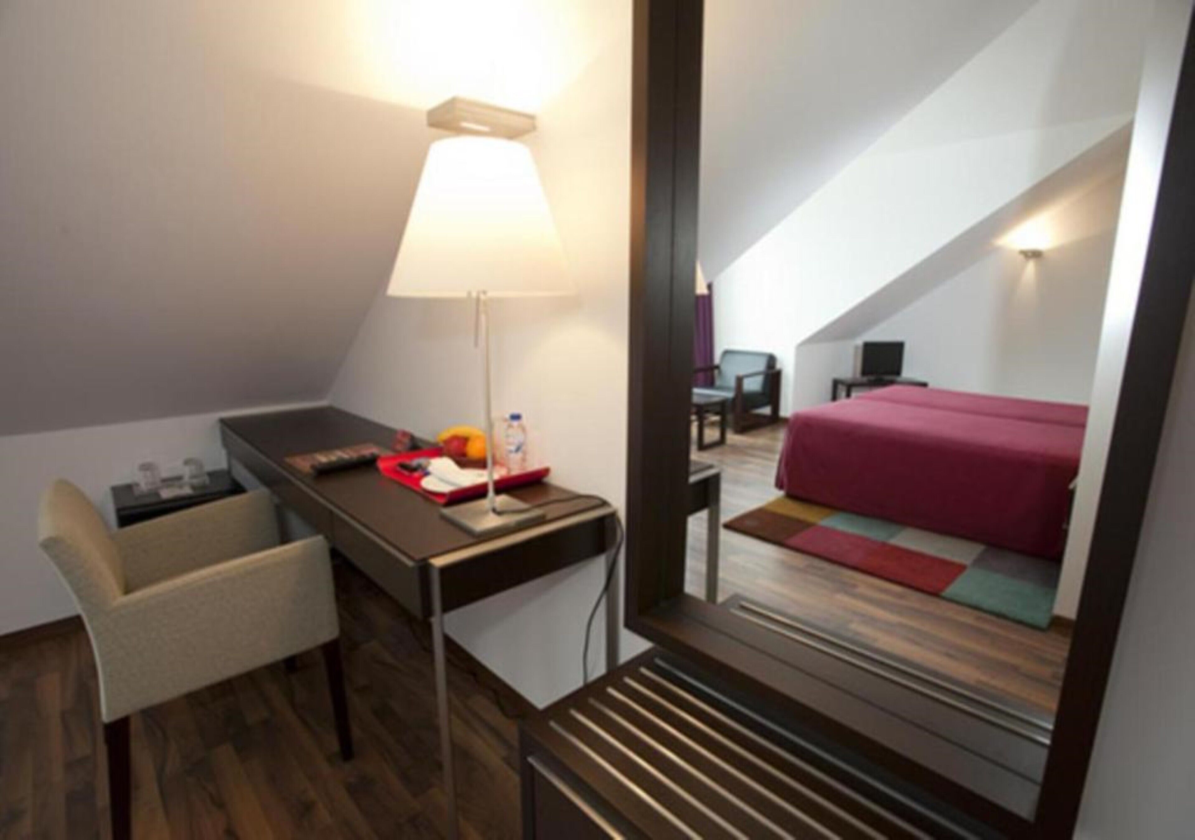 Una habitación de hotel moderna con escritorio, silla, lámpara y cama con colcha roja.