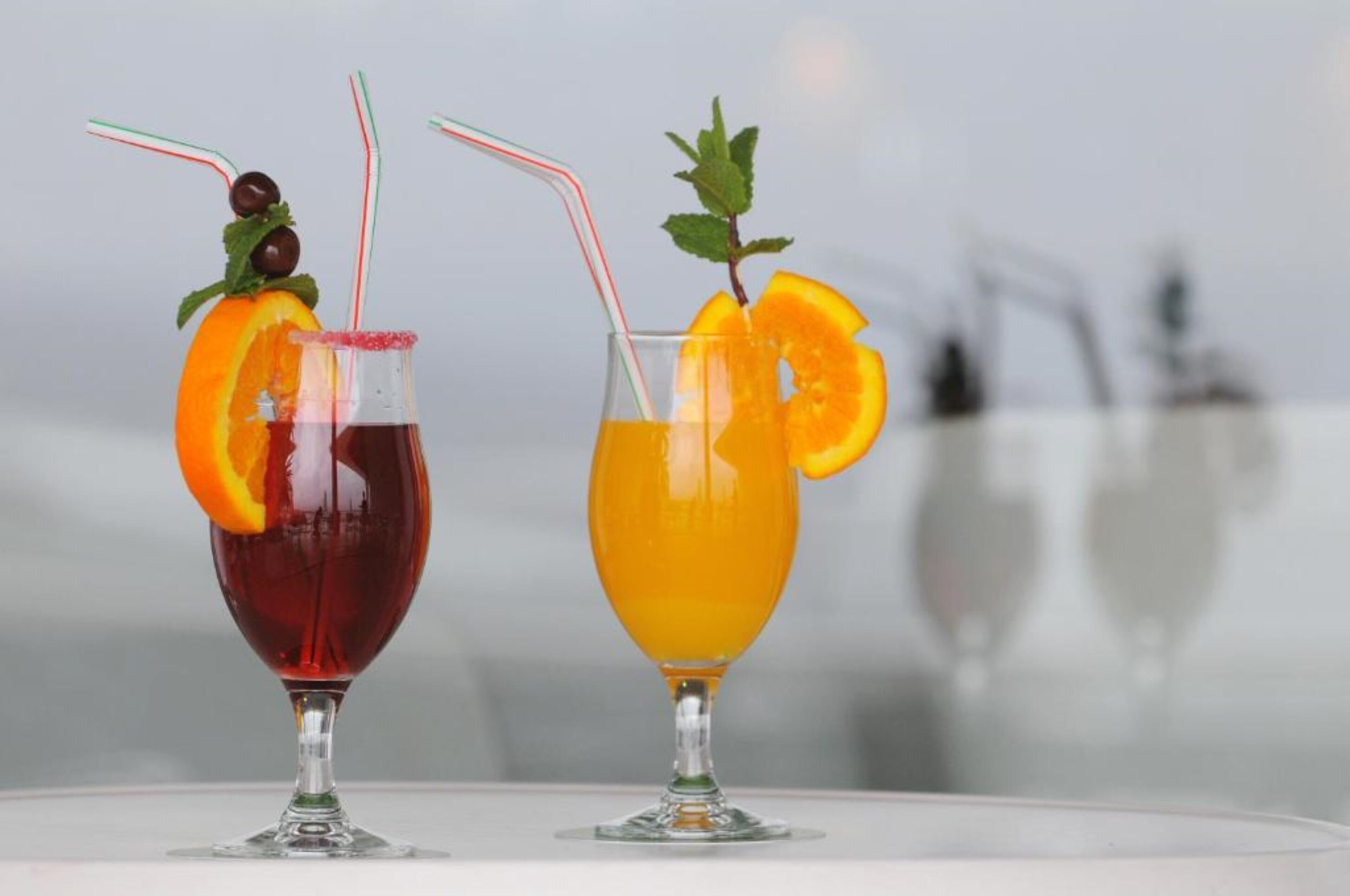 Deux cocktails servis dans des verres à pied, avec des pailles, et décorés de tranches d’orange et de feuilles de menthe.