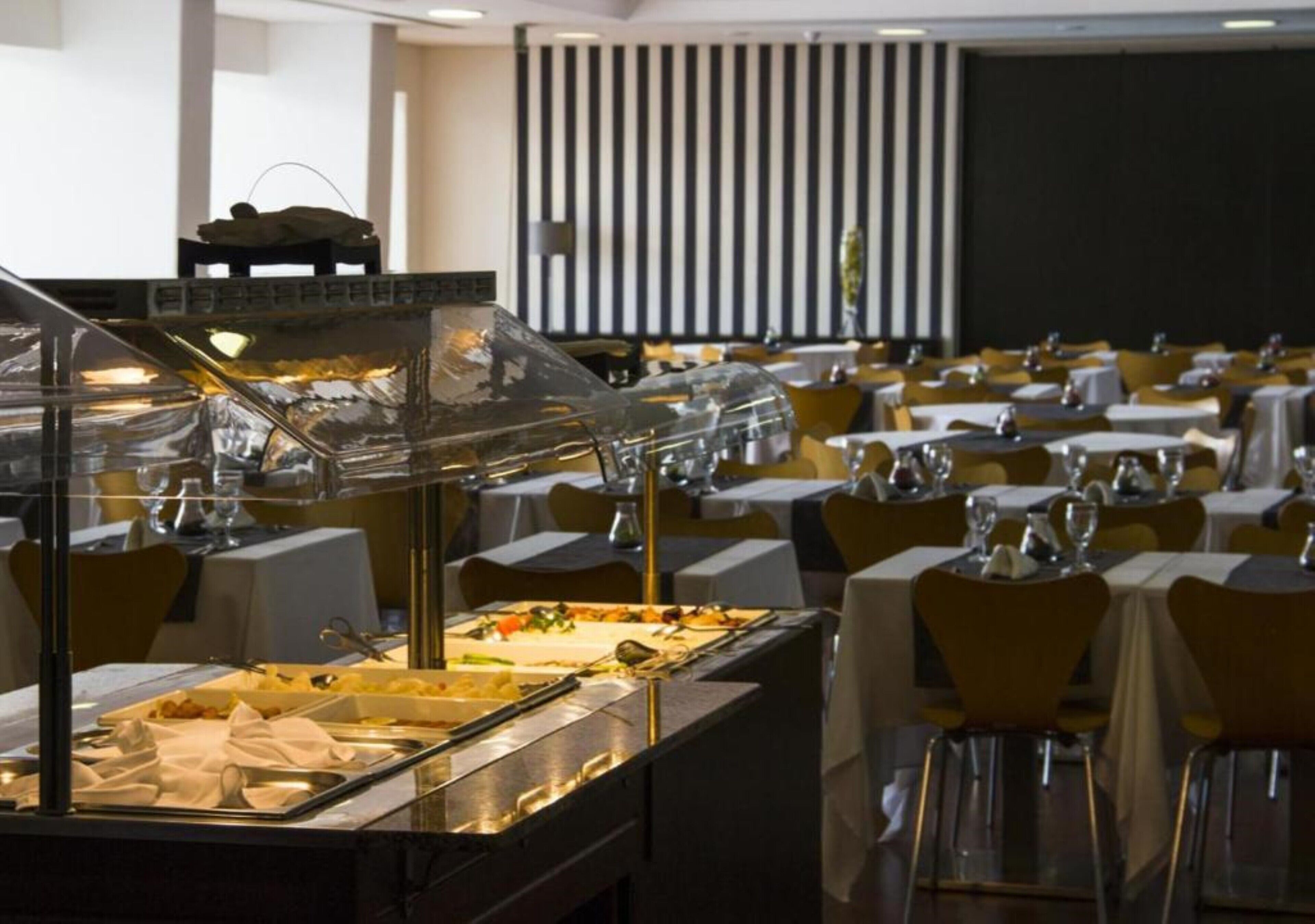 Disposición de un bufé con múltiples estaciones de servicio, mesas y sillas dispuestas en un amplio salón comedor.