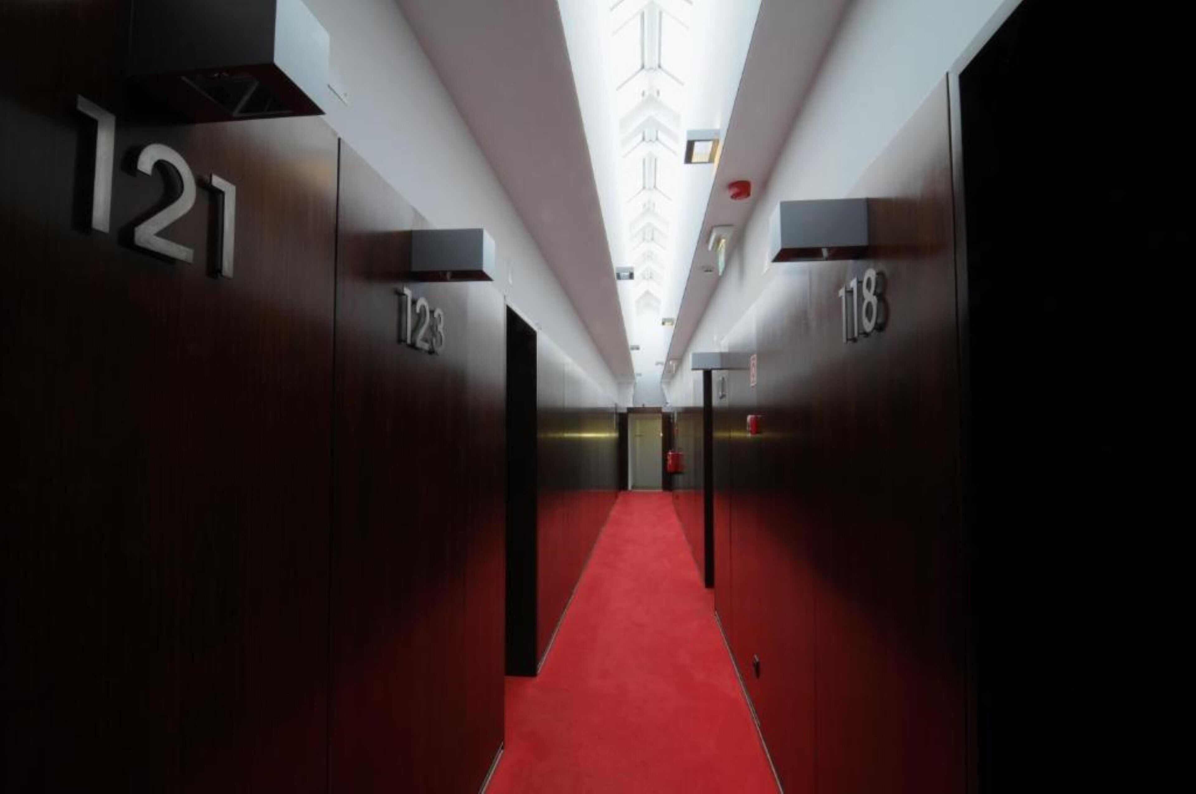 A modern hallway with red carpet, numbered doors, and a central light fixture.