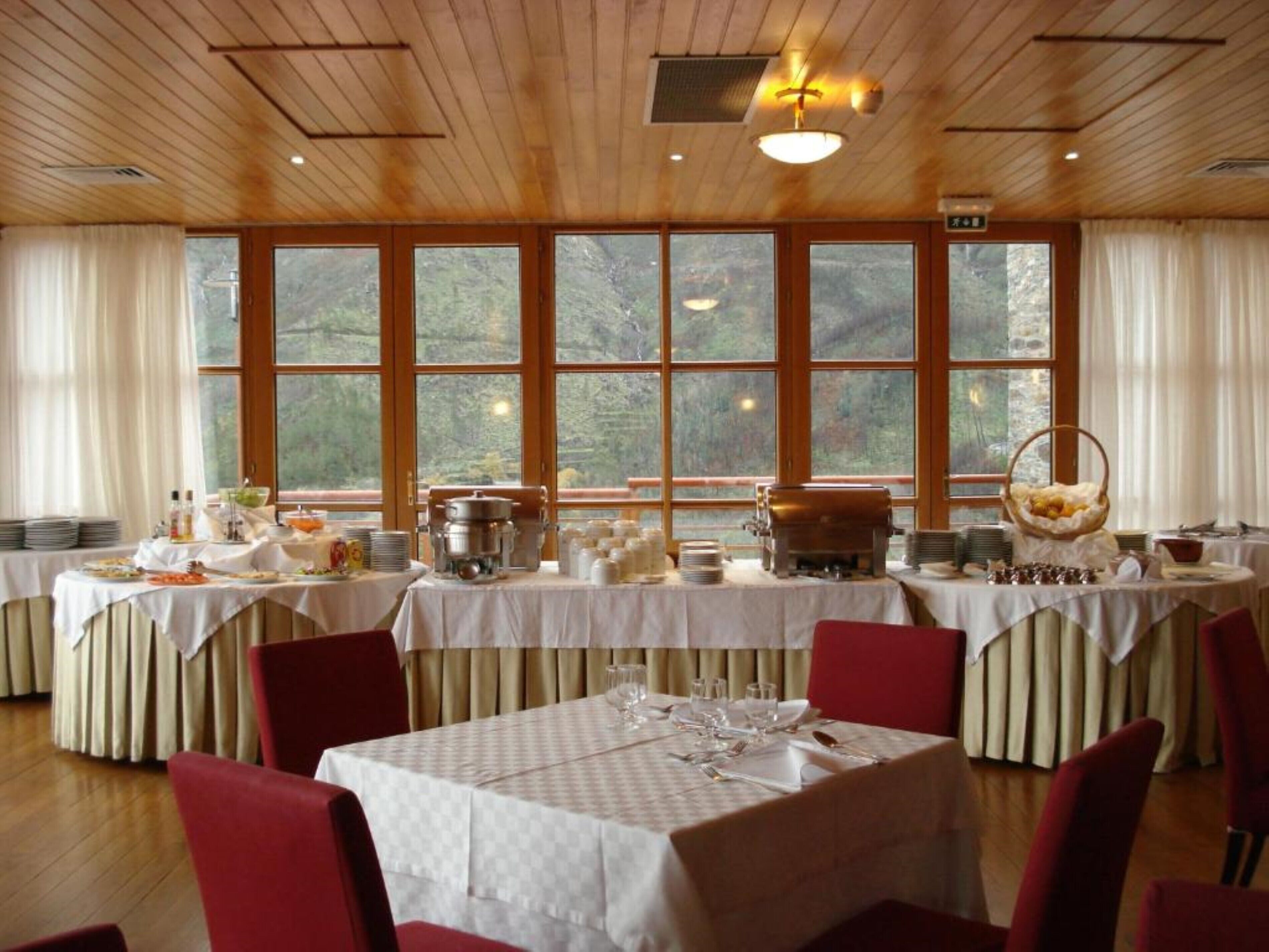 A dining area with multiple tables set for service, large windows offering a view of the outdoors, and a wooden ceiling.