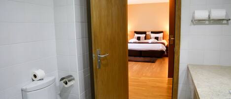 Et badeværelse med toilet, toiletpapir og en trædør, der fører til et soveværelse med seng og natbord.