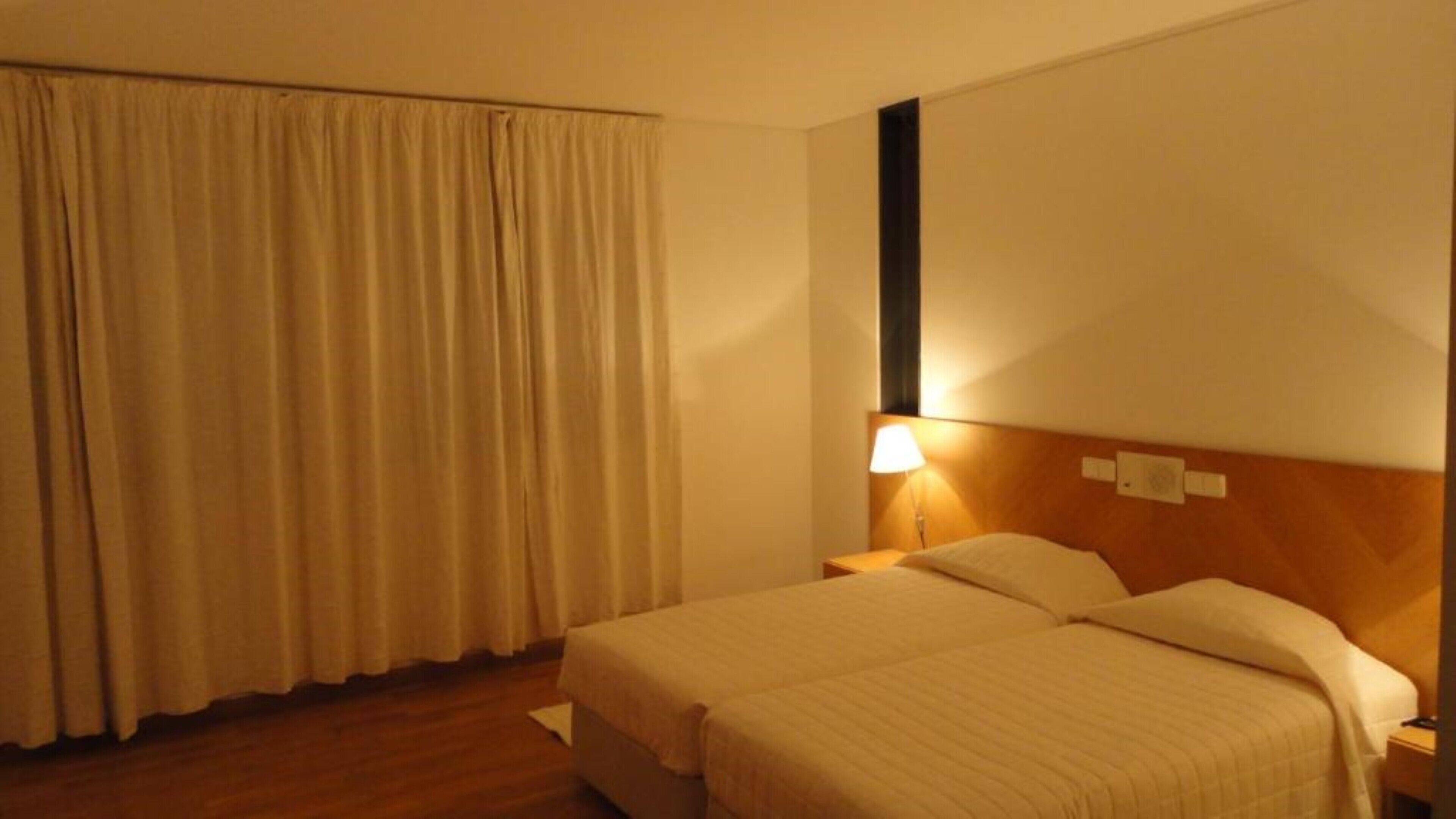 Una habitación de hotel con cama, mesita de noche, lámpara y ventana con cortinas.