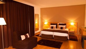 A hotel room with a large bed, two bedside lamps, a chair, a small table, and a mirror. - INATEL Caparica (Almada)