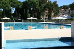 A swimming pool with numbered lanes, surrounded by trees and umbrellas, with buildings in the background. - INATEL Caparica (Almada)