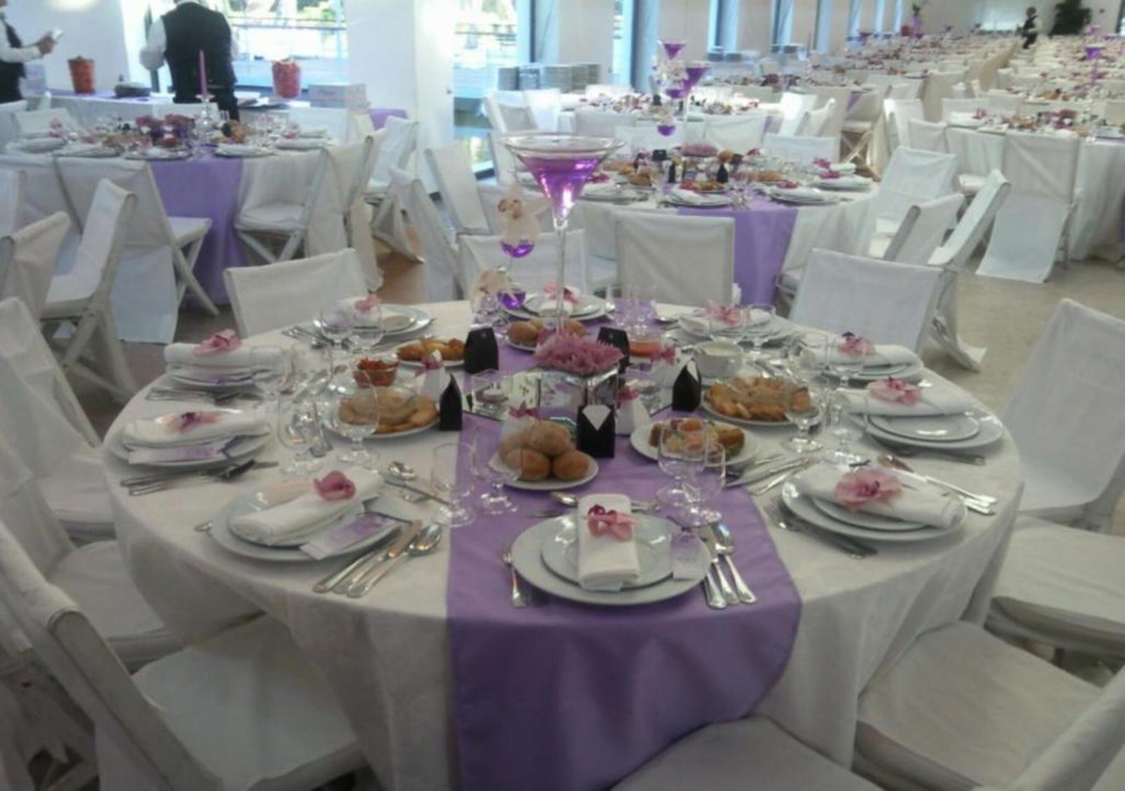 A banquet hall with round tables covered in white tablecloths and purple centerpieces, arranged in rows with white chairs.