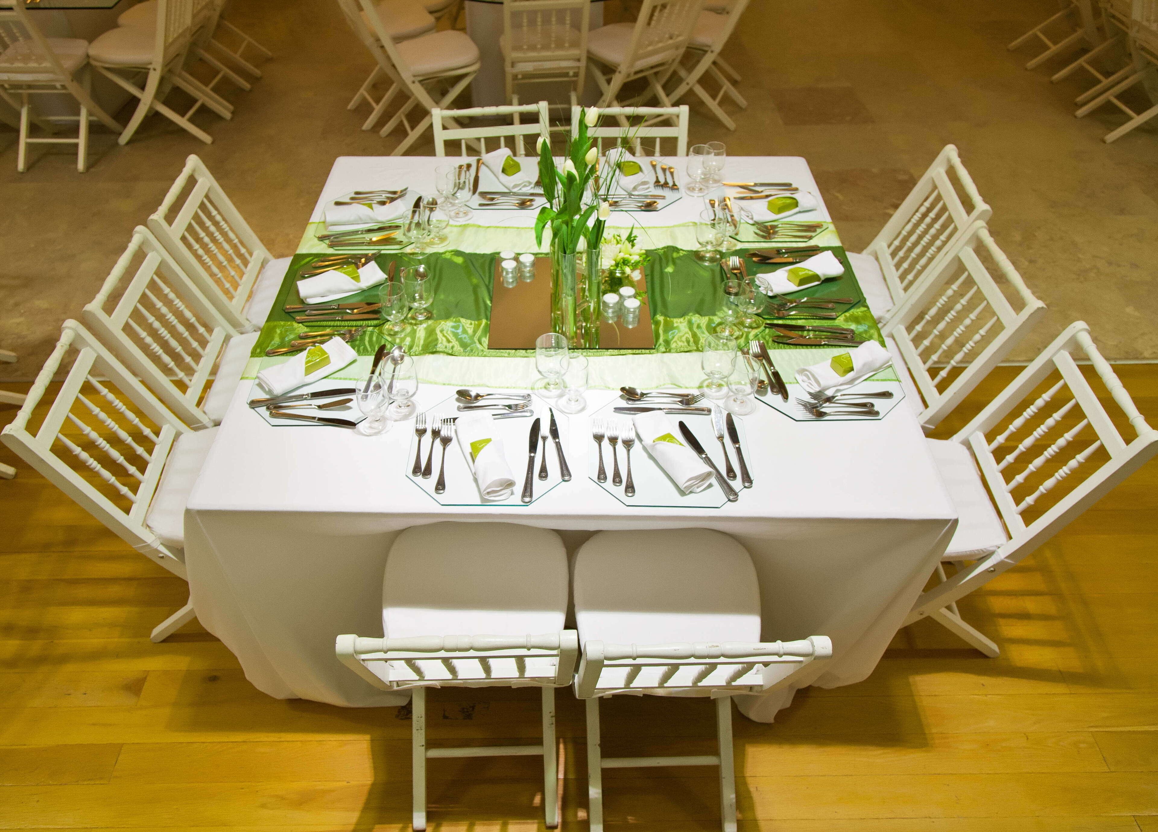 A round dining table set for six with white chairs, green table runners, and silverware.