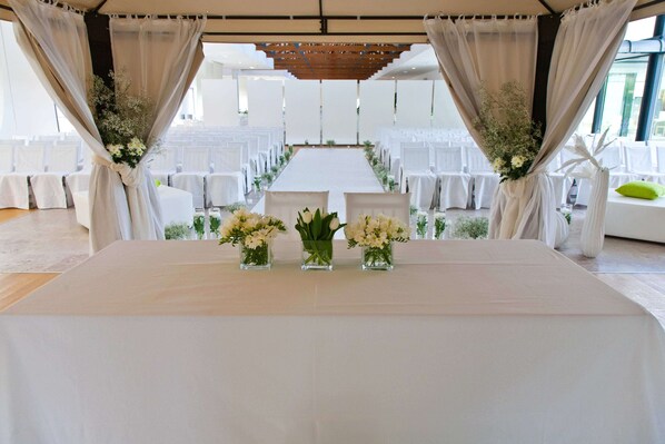 An indoor venue with a long table at the front, flanked by white chairs and draped curtains.