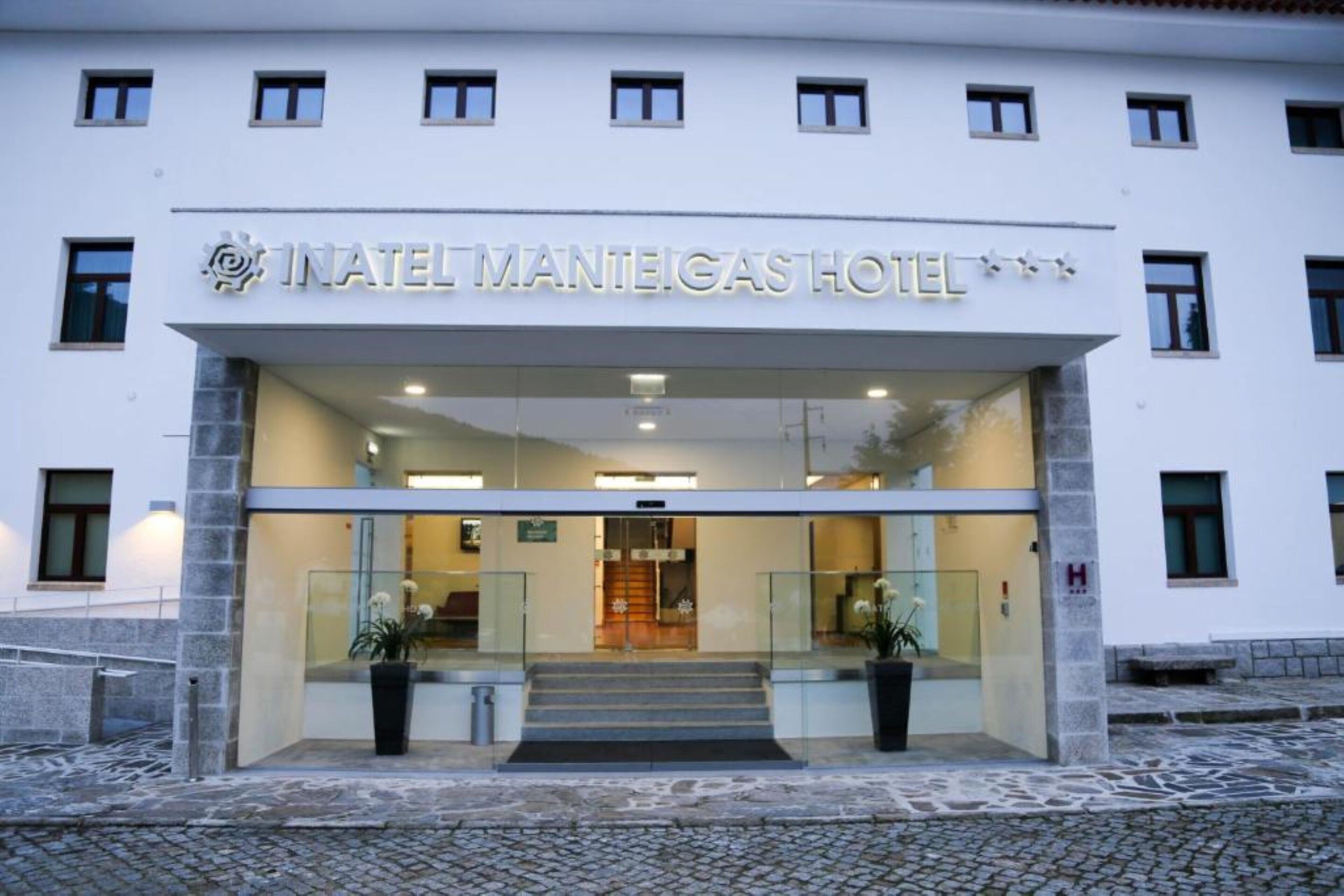 The entrance to the NATEL MANIEGAS HOTEL with a glass facade, a staircase leading to the lobby, and a clear view of the interior.