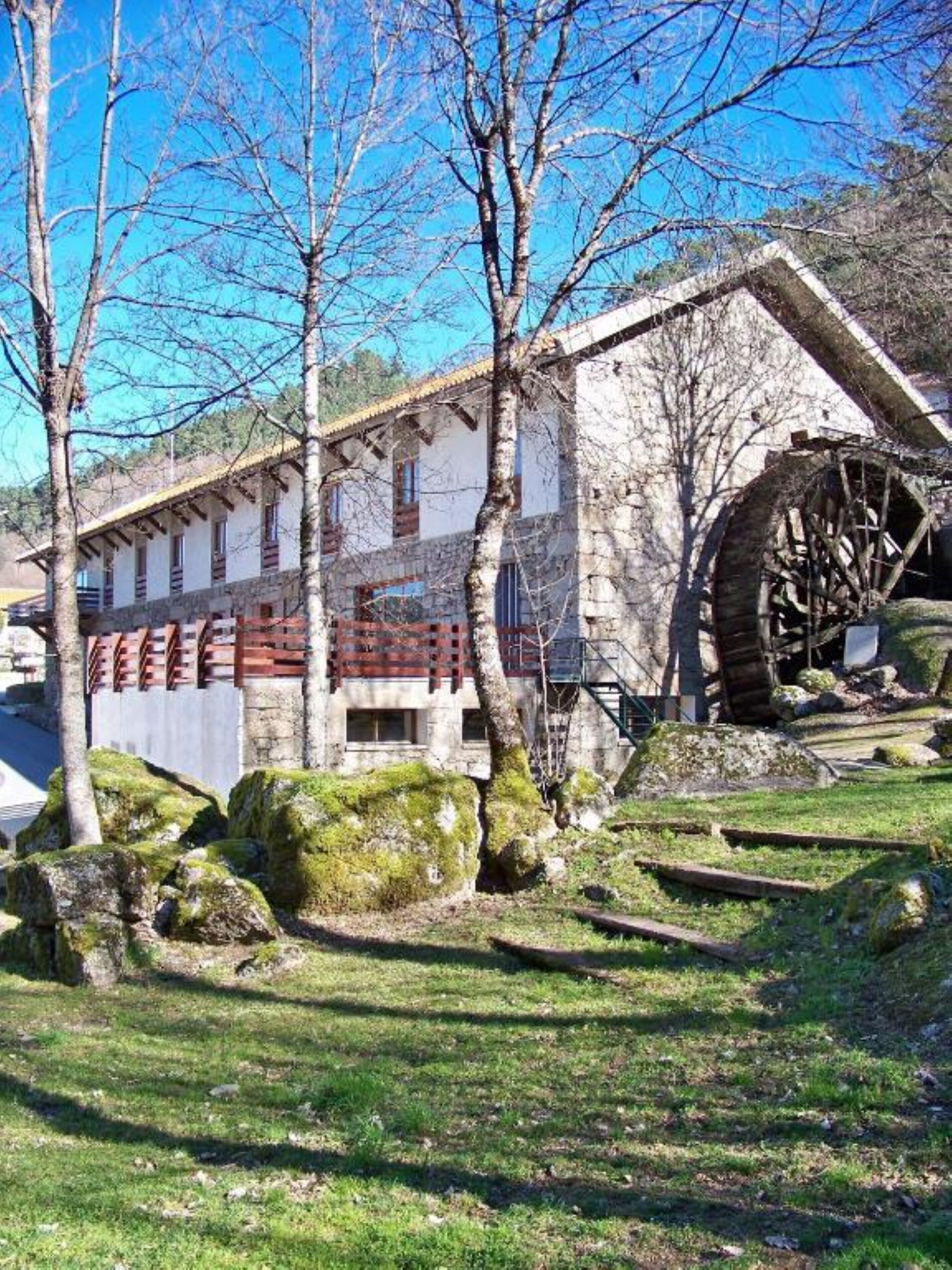 A rustic building with a large water wheel, surrounded by trees and rocks.