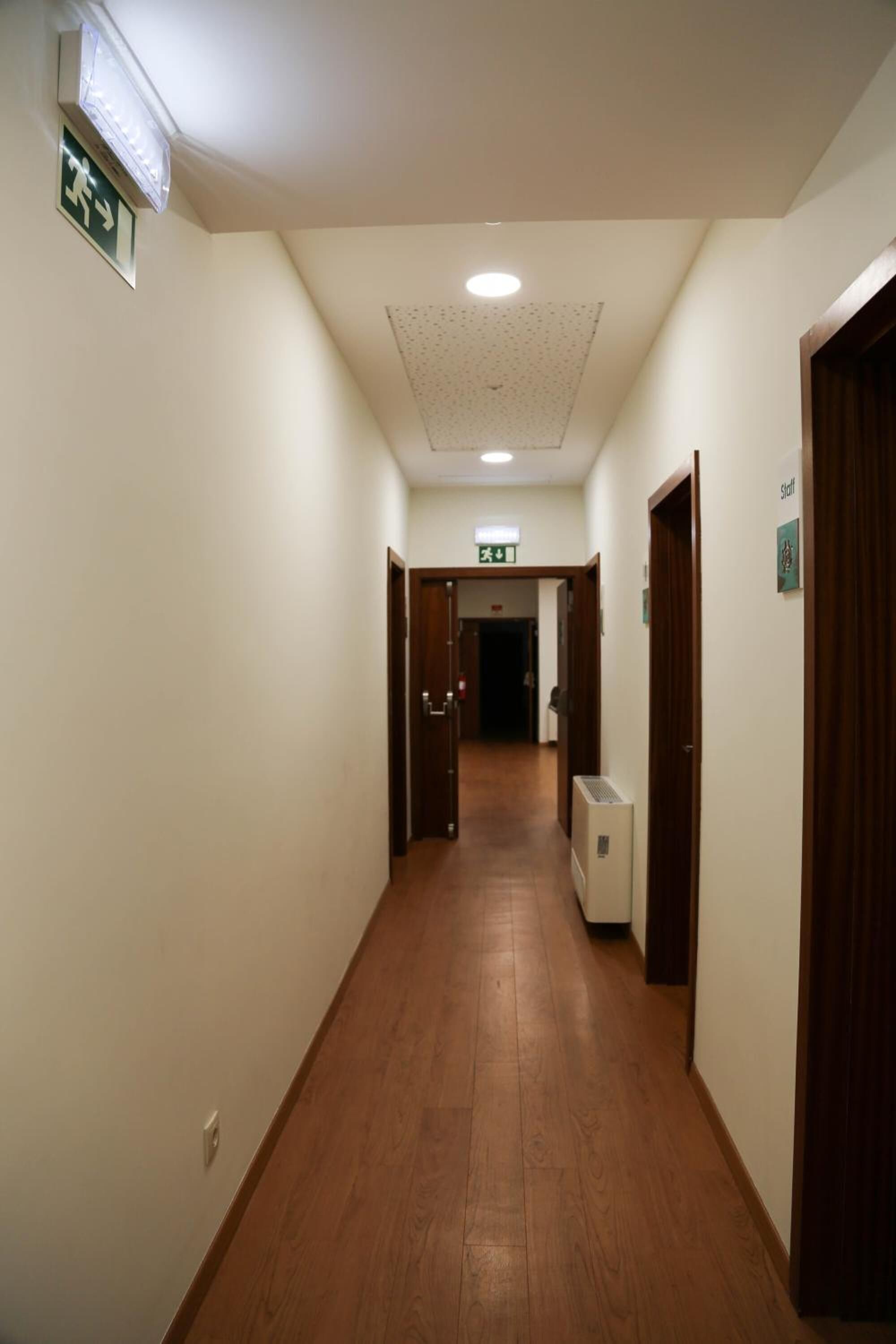 A narrow, well-lit hallway with wooden flooring, doors on both sides, and an exit sign on the left.