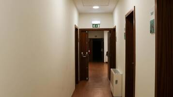 A narrow, well-lit hallway with wooden flooring, doors on both sides, and an exit sign on the left.