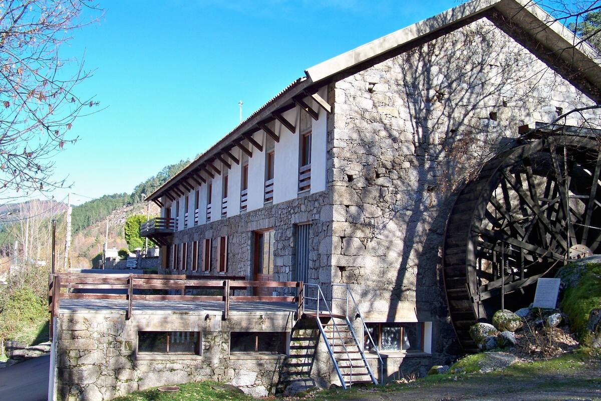 A stone building with a large water wheel attached to its side, a wooden staircase leading to a balcony, and a view of a road and hills in the background.