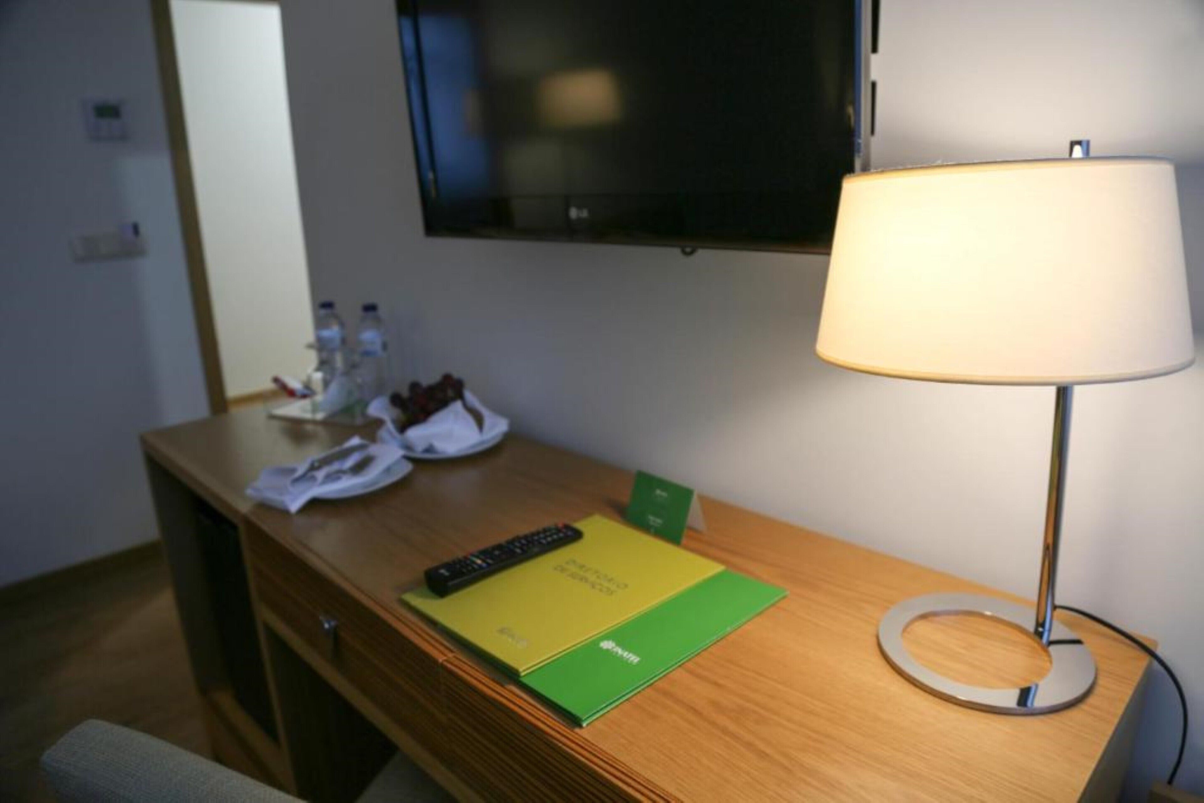 A hotel room with a wooden desk, a lamp, a television, and a green folder on the desk.