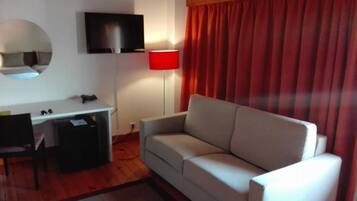 Une pièce comprenant un bureau, un téléviseur fixé au mur, une lampe rouge, un canapé gris et une fenêtre avec des rideaux rouges.