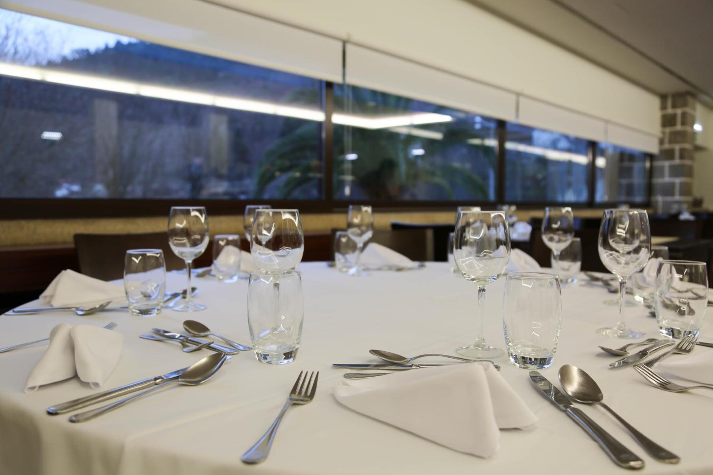 Una mesa de comedor dispuesta con cubiertos y vasos transparentes, con vistas a árboles a través de la ventana.