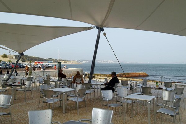 An outdoor café with tables and chairs, overlooking a beach and the sea. - INATEL Oeiras (Oeiras)