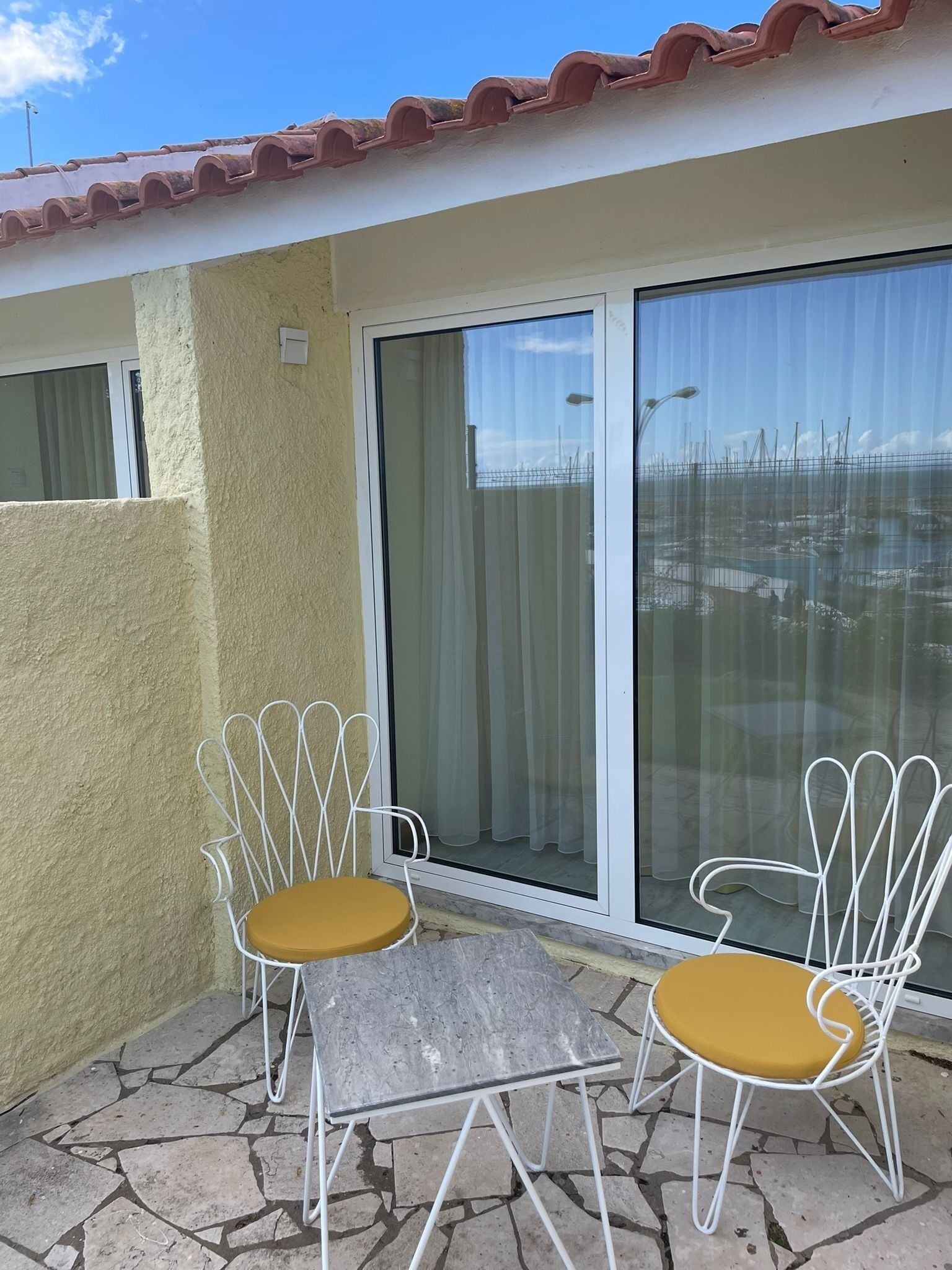 A patio with two white metal chairs with yellow cushions, a small table, and a sliding glass door leading to a covered area.