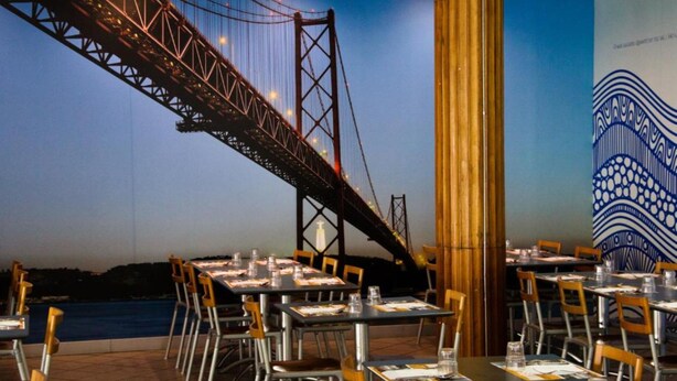 A restaurant with tables set for dining, a view of a bridge, and a mural on the wall. - INATEL Oeiras (Oeiras)