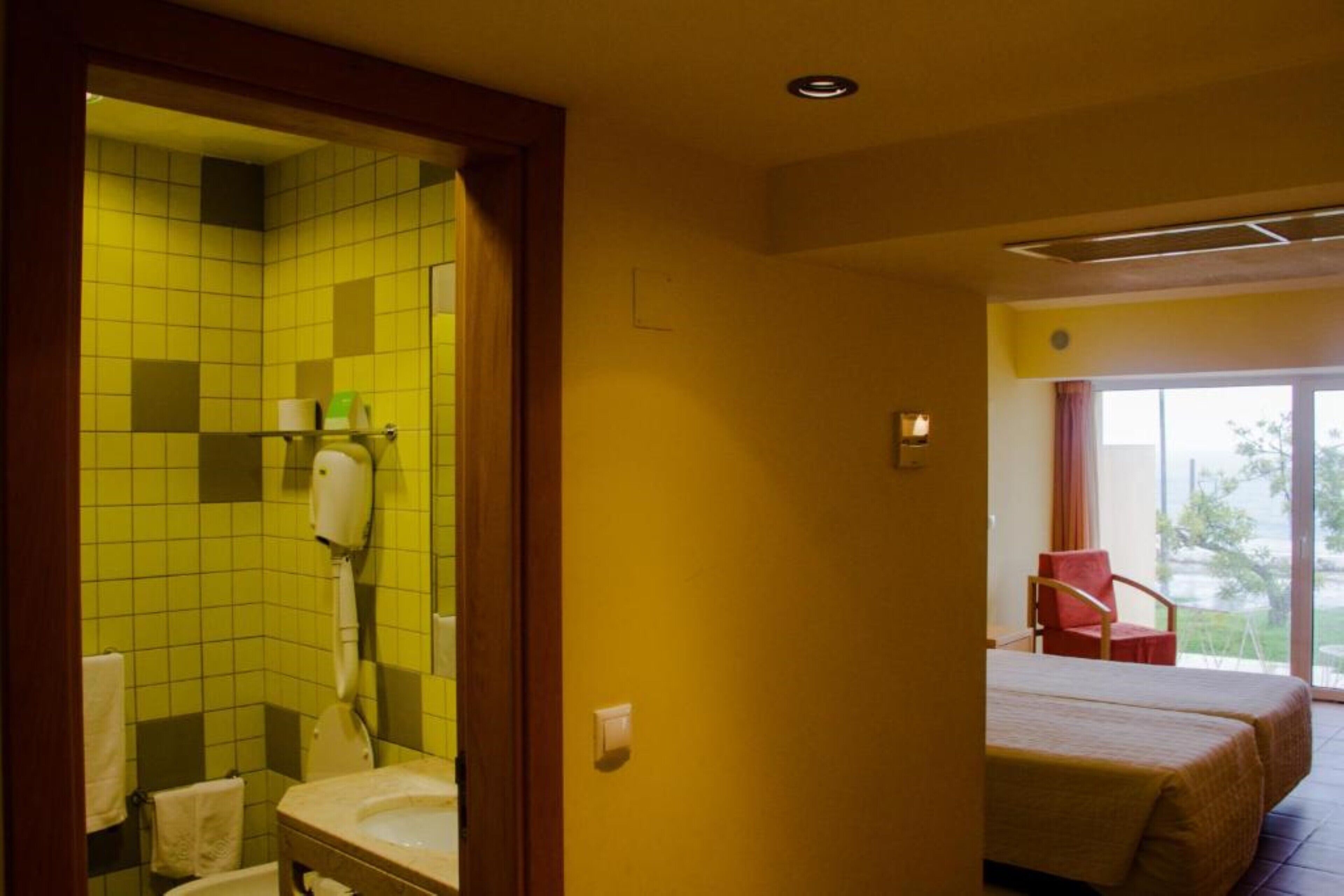 A hotel room with a bathroom featuring yellow tiles, a sink, and a toilet. There is a bed with a red chair and a window with a view.