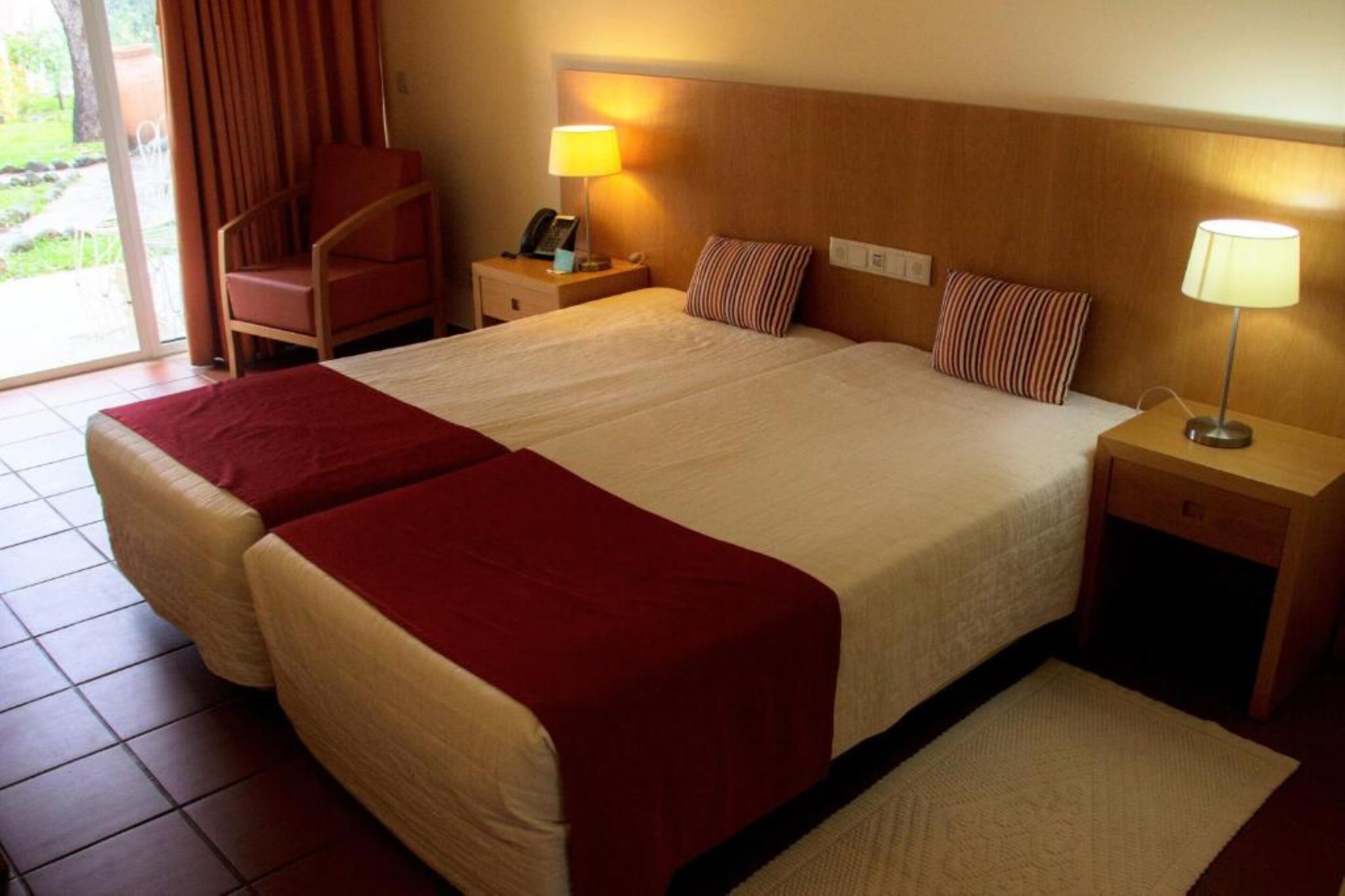 A double bed with a wooden headboard, two bedside tables with lamps, a red chair, and a view of the outdoors through a sliding door.