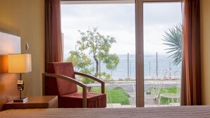 A hotel room with a large bed, a chair, a nightstand, and a view of the beach and ocean through a sliding glass door. - INATEL Oeiras (Oeiras)