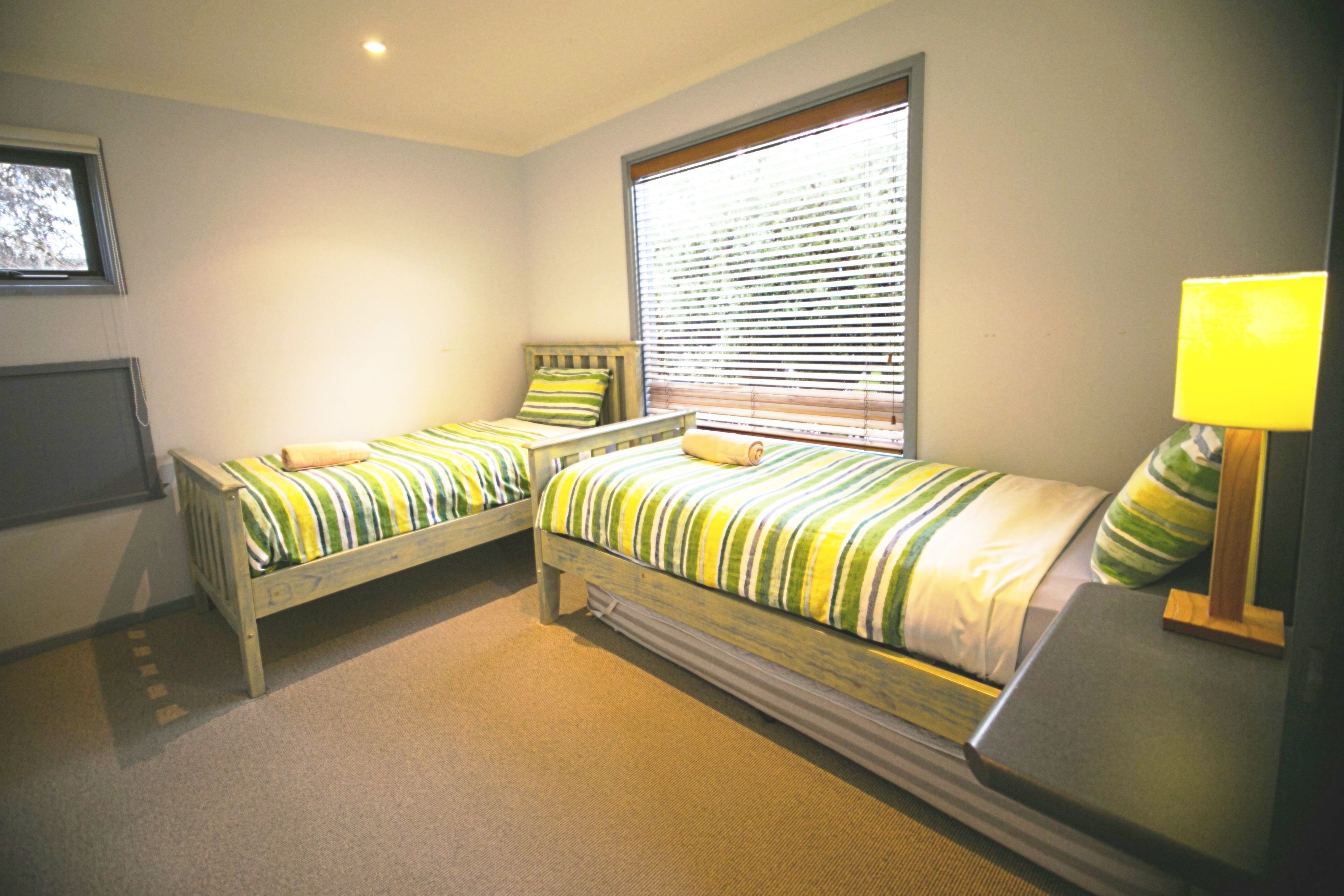 A room with two single beds, a window with blinds, a bedside table with a lamp, and a wall-mounted TV.