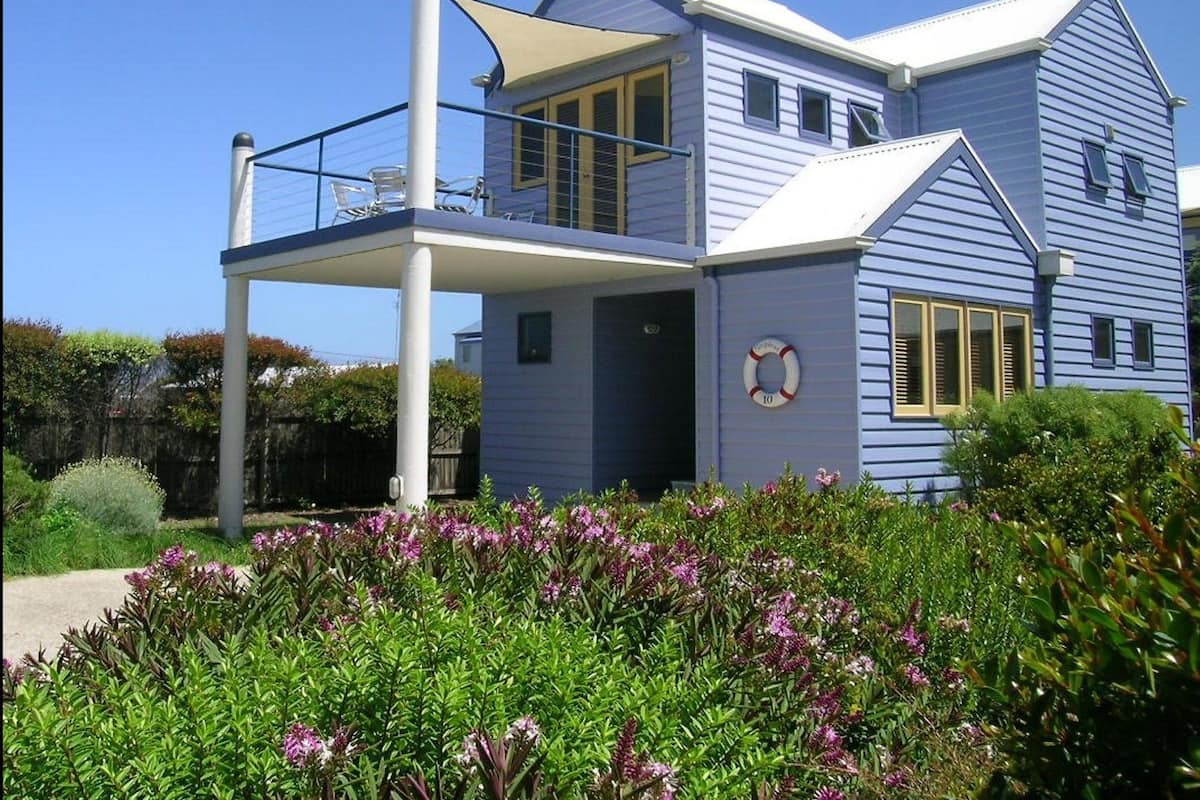 A two-story house with a balcony, a lifebuoy on the wall, and a garden with purple flowers.