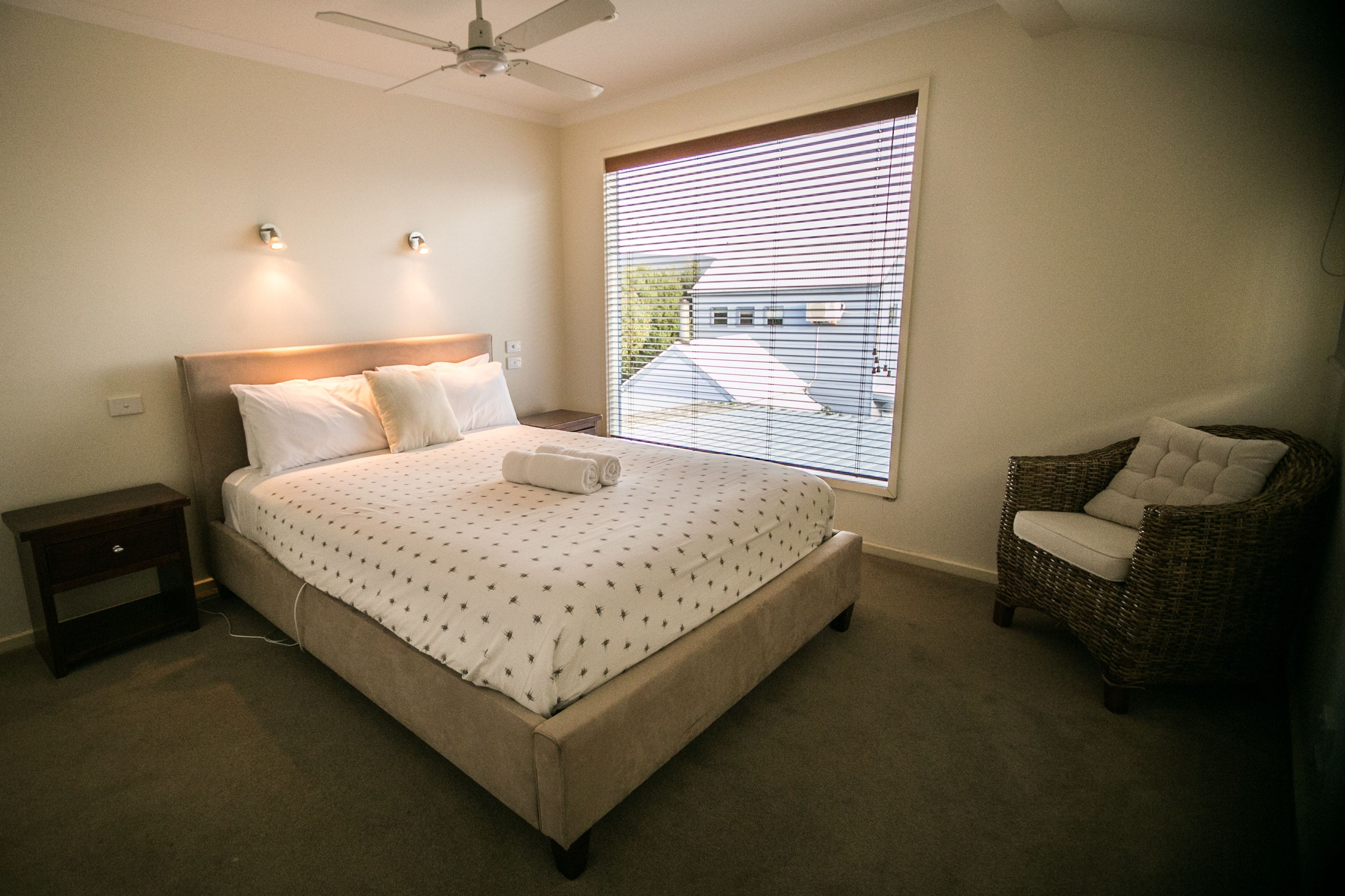 Quarto com cama grande, mesa de cabeceira, cadeira, ventilador de teto e janela com persianas.