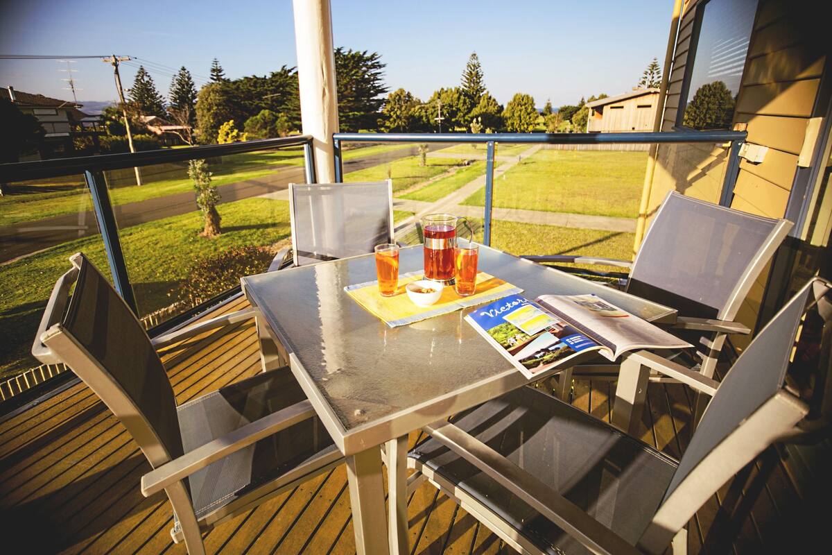A table with chairs on a balcony with a view of a grassy area and houses.