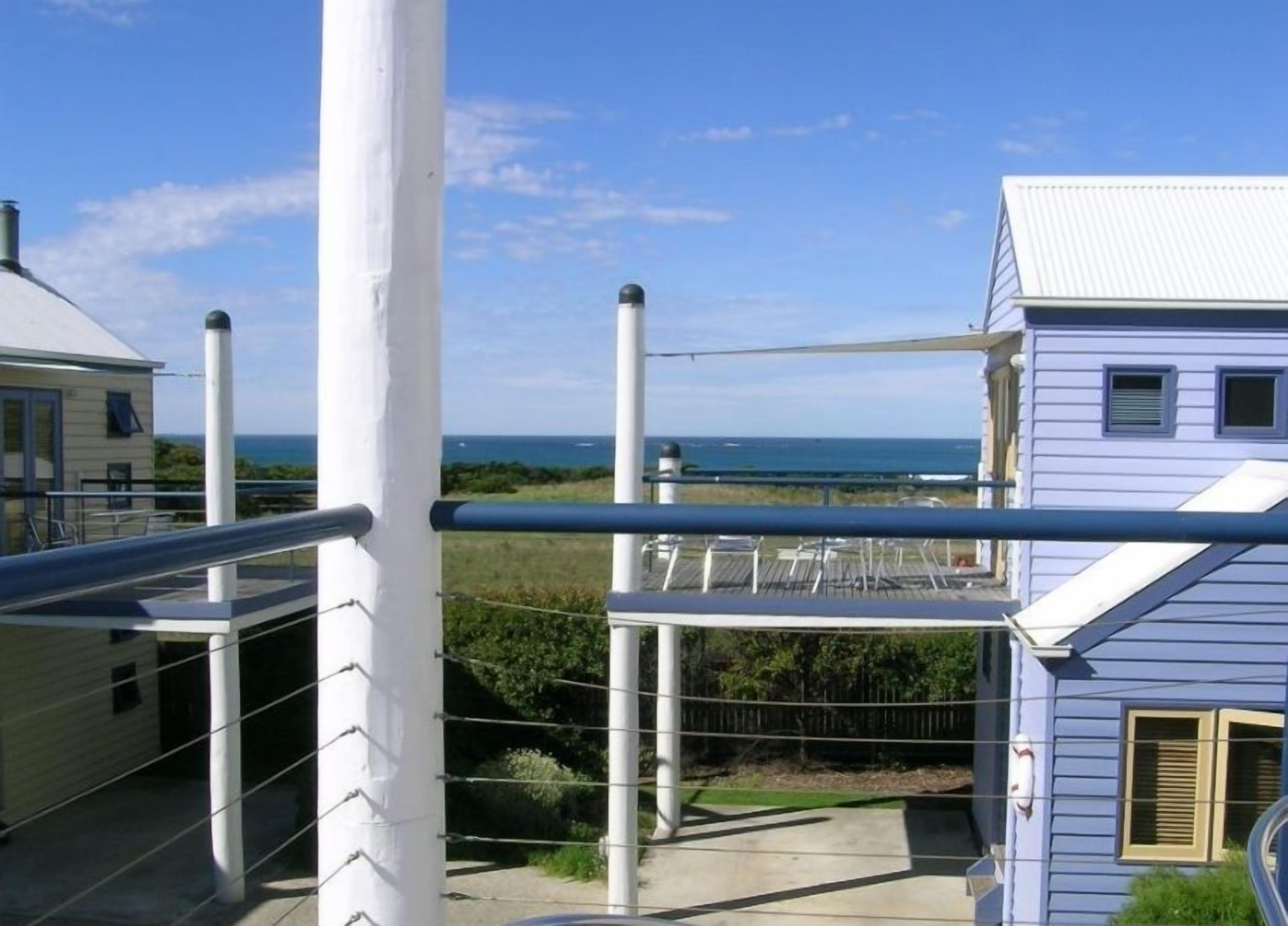 Une maison située sur la côte, dotée d’un balcon donnant sur la mer, d’une clôture blanche et surplombée d’un ciel dégagé.