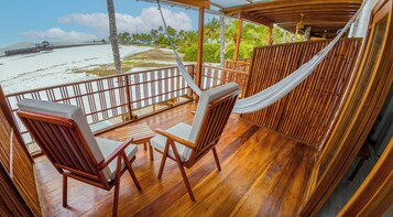 Una terraza de madera con dos sillas y una hamaca, con vista a una playa con palmeras y cielo despejado.