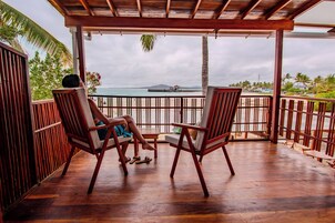 Una terraza de madera con dos sillas mirando al mar, una barandilla de madera y una palmera.