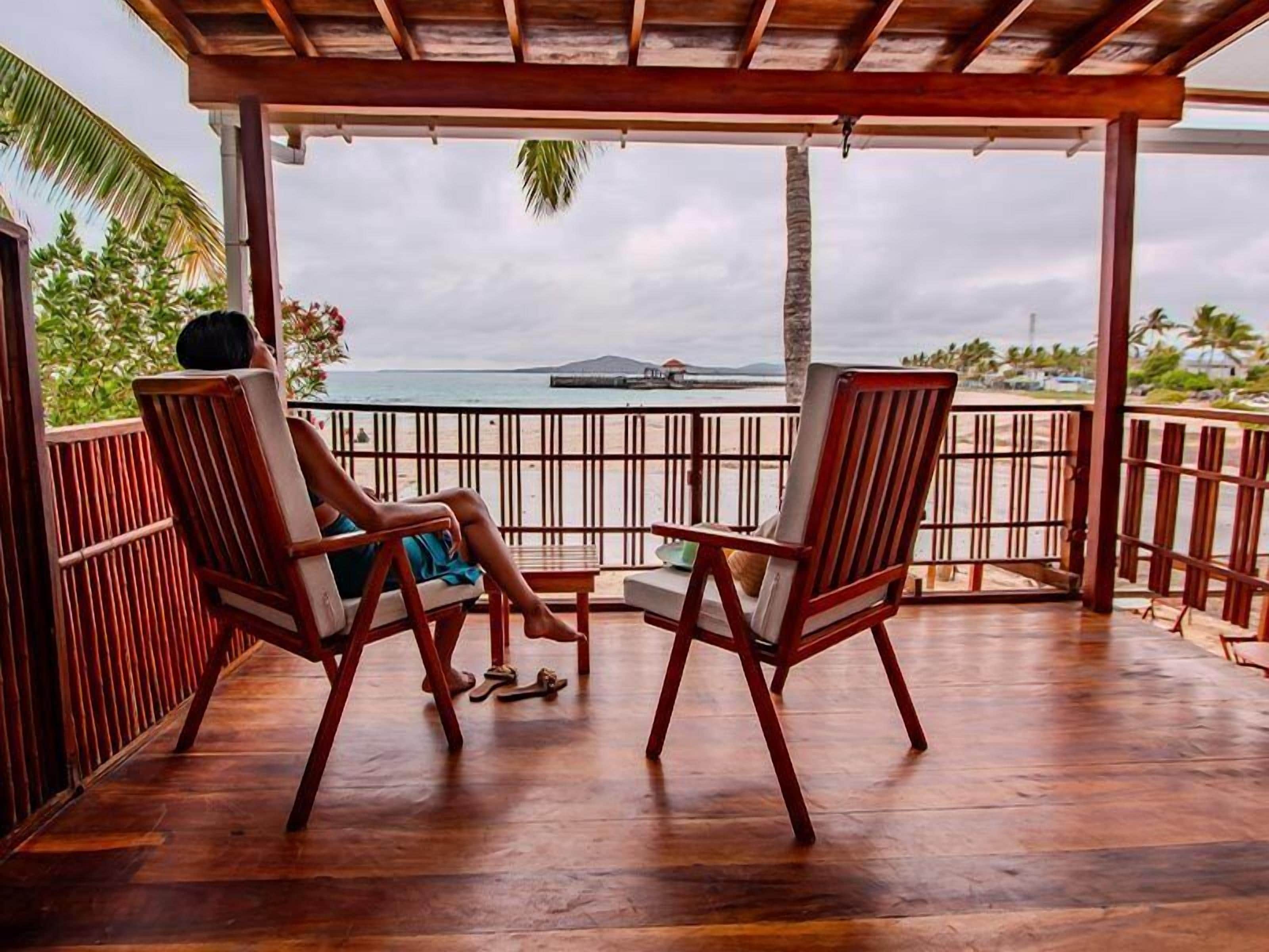 A wooden deck with two chairs facing a beach view, a wooden railing, and a palm tree.