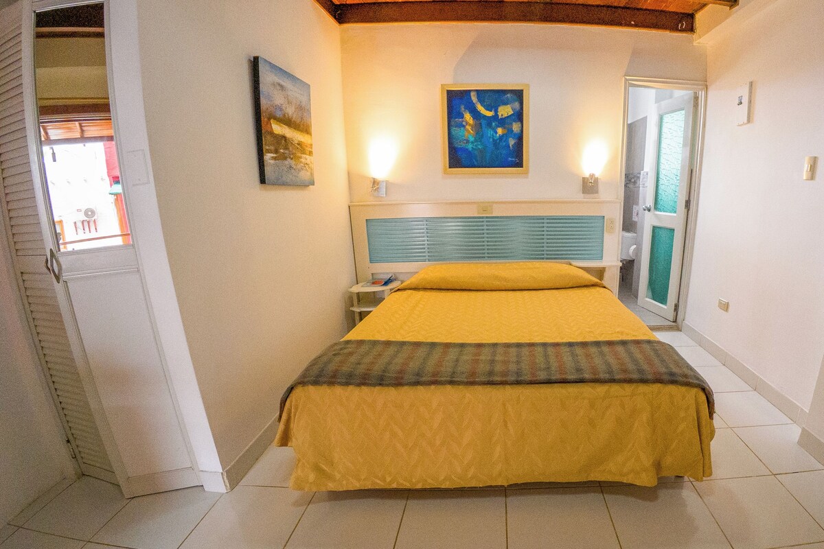 Habitación de hotel con cama, mesita de noche, un cuadro y un baño visible a través de una puerta abierta.