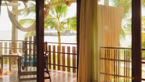 Una terraza de madera con una hamaca, una mesa y sillas, con vista a una playa con palmeras.