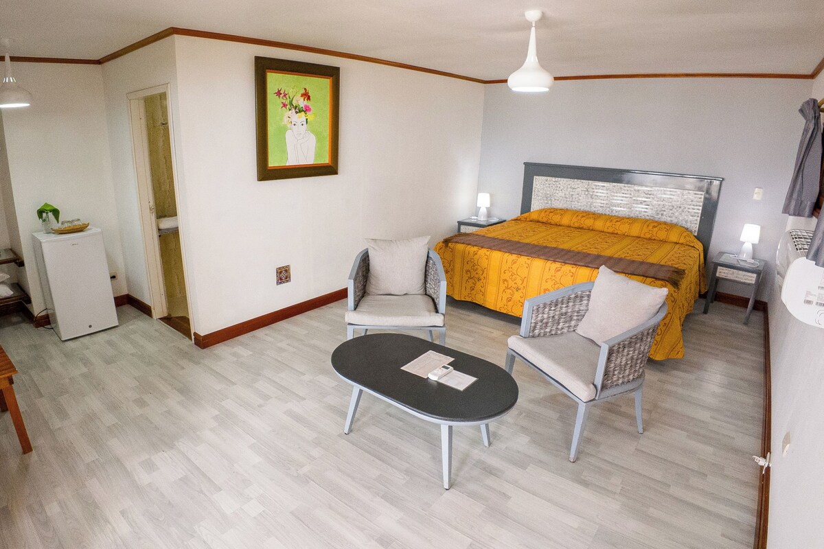 A hotel room with a bed, two chairs, a small table, a refrigerator, and a painting on the wall.