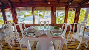 Un comedor con mesa de vidrio y sillas blancas, con vistas a una playa con palmeras.