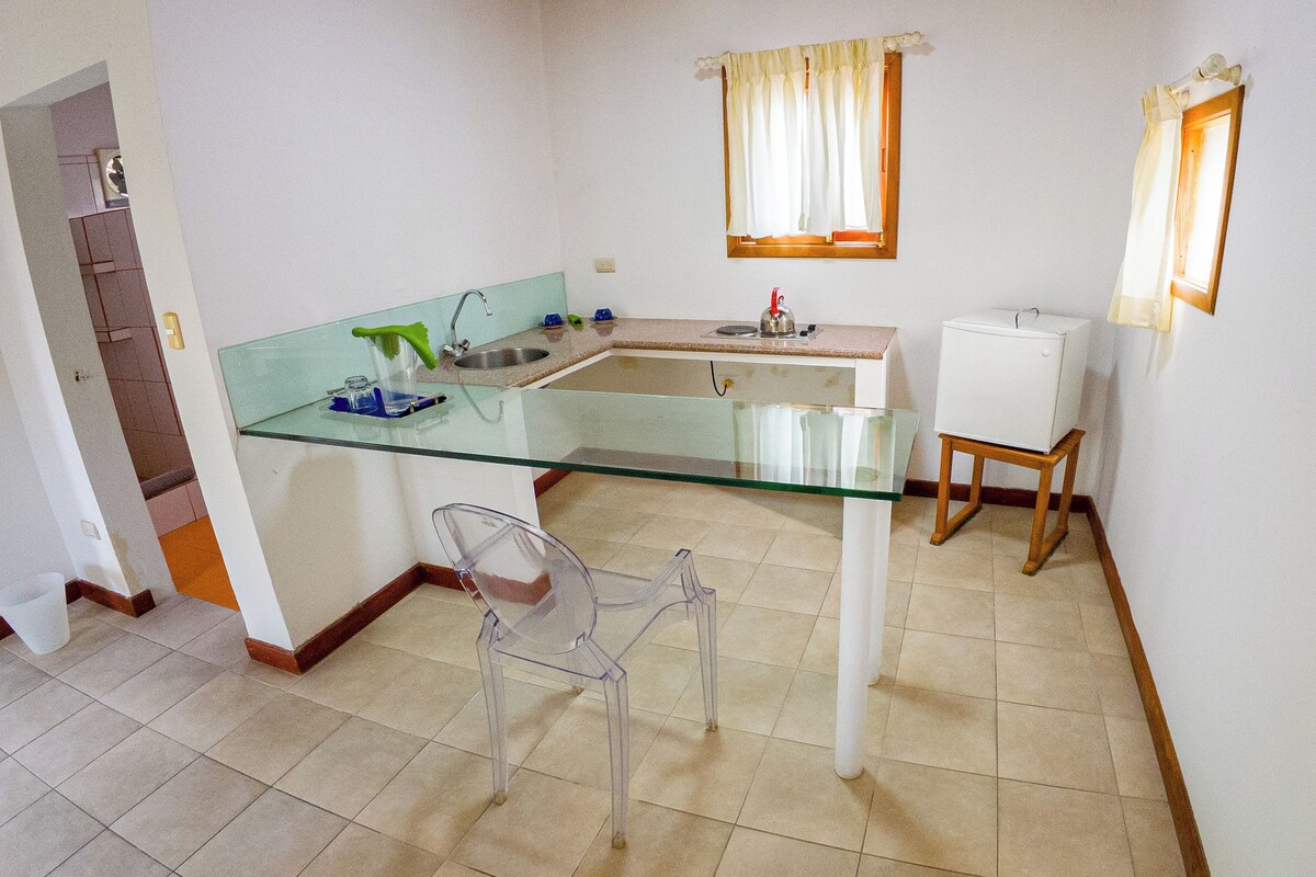 A small kitchen with a glass countertop, a sink, a refrigerator, and a chair.