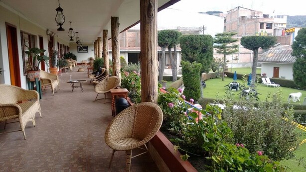 Un espacio al aire libre techado con muebles de mimbre, plantas en macetas y vistas a un jardín cuidado y edificios cercanos.
