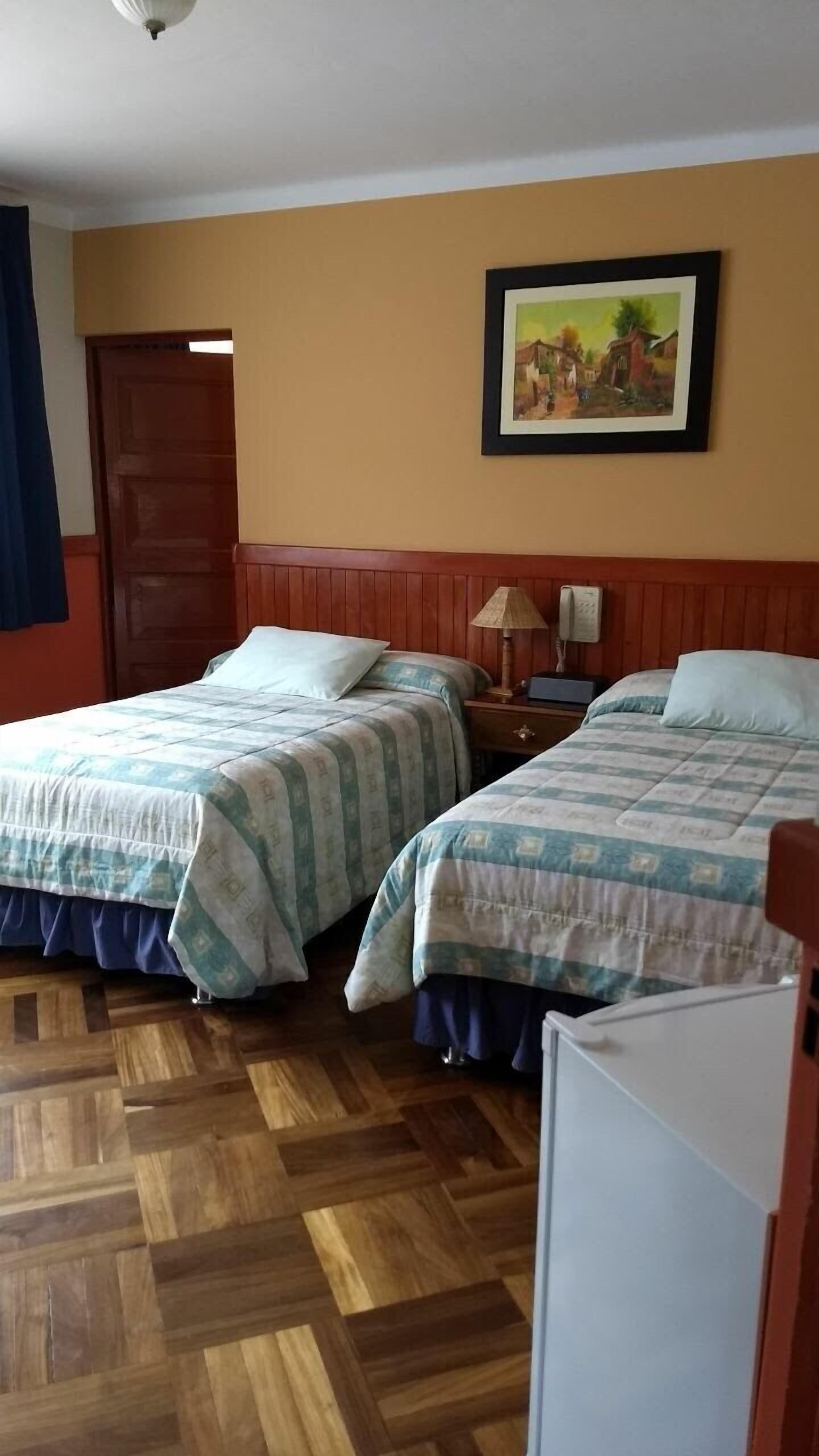 A hotel room with two beds, wooden flooring, a painting on the wall, and a door in the background.