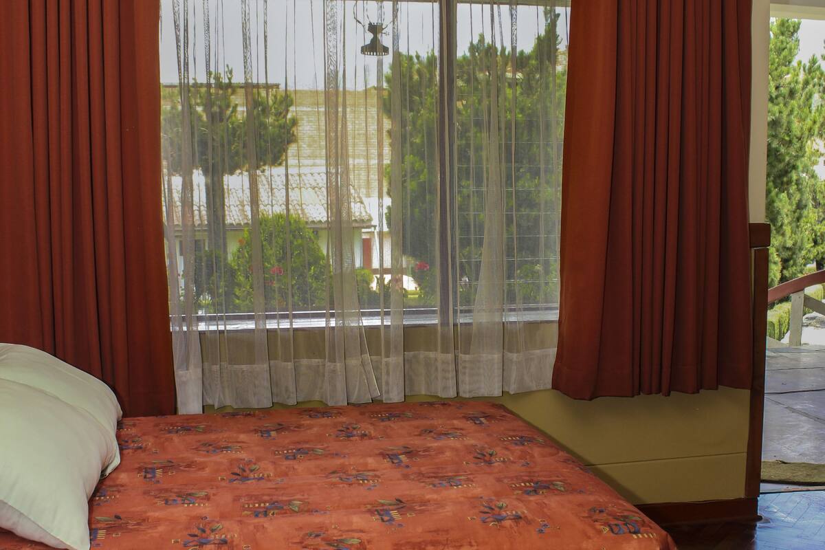 A bedroom with a large bed, a window with sheer curtains, and a view of greenery outside.
