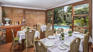 A dining area with wicker chairs and tables set for a meal, overlooking a swimming pool and lush garden. - Algoa Guesthouse Summerstrand (Port Elizabeth)