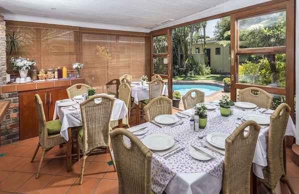 Comedor con sillas y mesas de mimbre dispuestas para una comida, con vista a una piscina y un jardín exuberante.